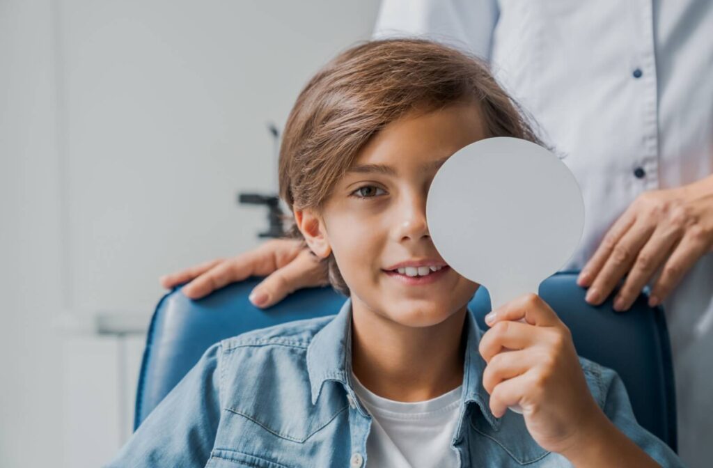 Smiling child sitting in an eye exam chair, holding an occluder over one eye while an optometrist stands behind them.