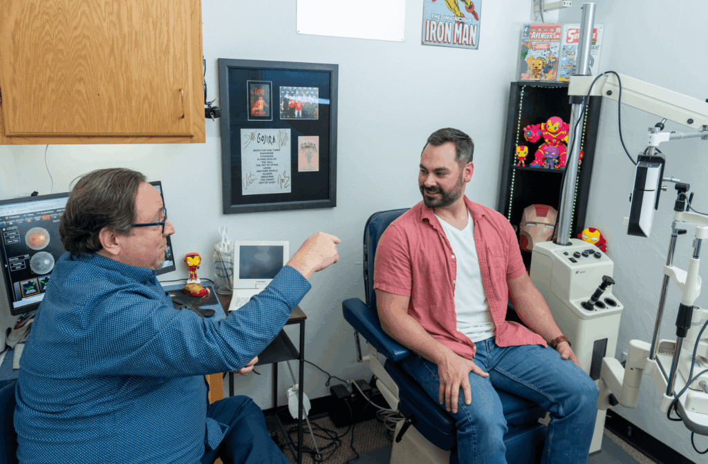 Optometrist speaking with a patient in an exam chair inside a decorated eye care office, with eye exam equipment and Iron Man-themed collectibles in the background.