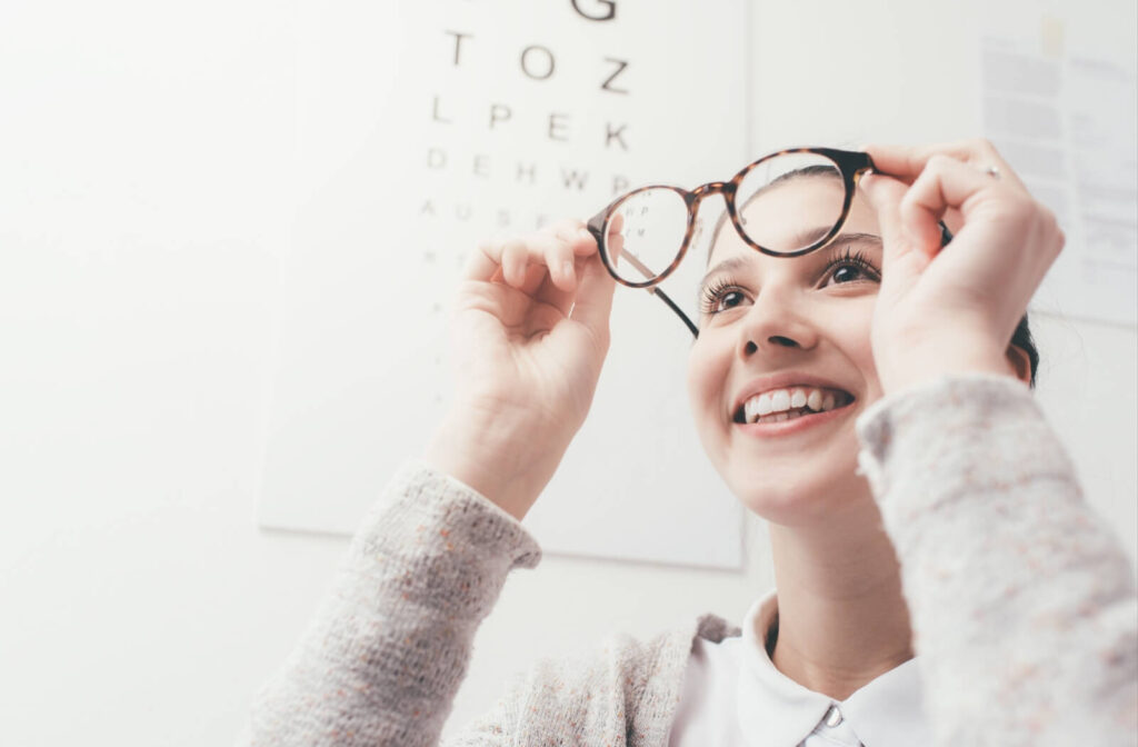 Smiling young woman trying on a pair of eyeglasses at an eye clinic, with an eye chart blurred in the background.