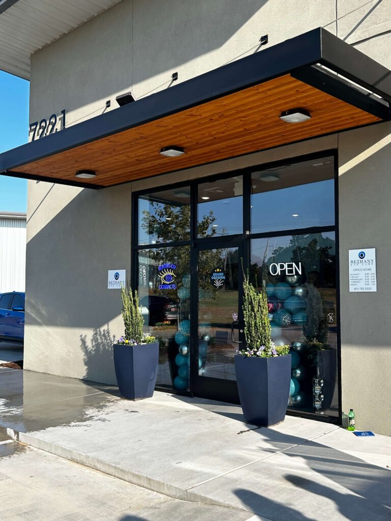 Exterior of an optometry clinic with large glass doors, blue planters, and “OPEN” and “Grand Opening” signs visible, reflecting a sunny day outside.