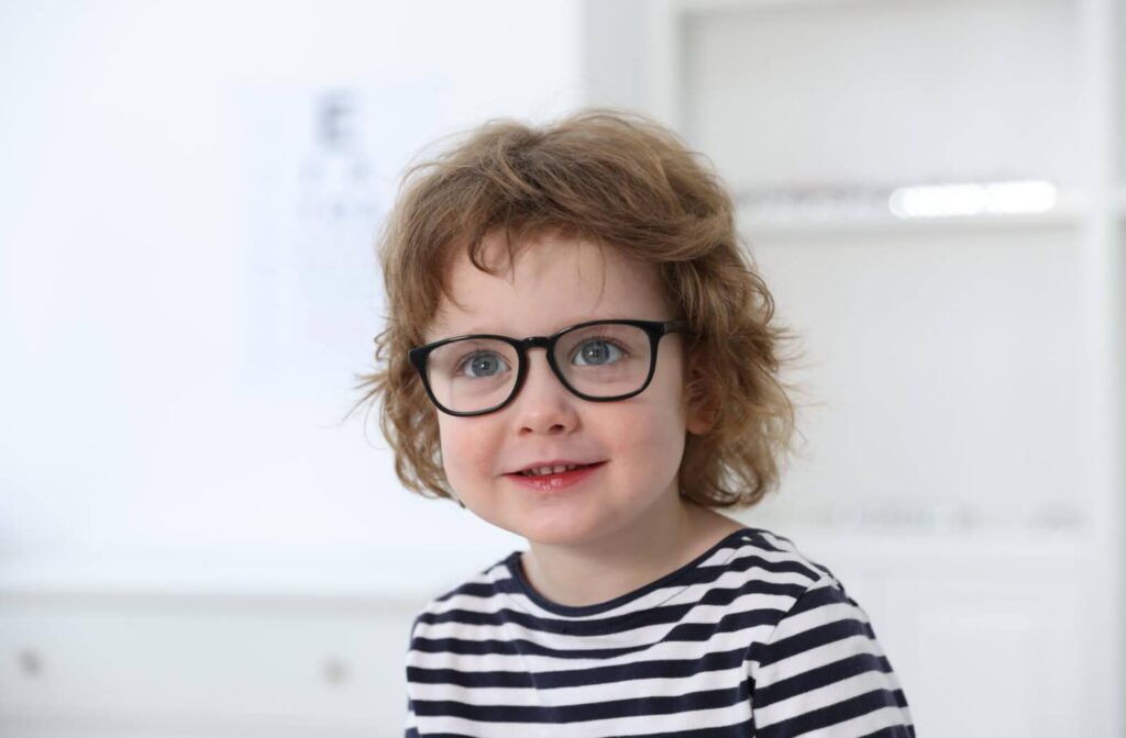 A smiling young boy with curly hair wearing black-rimmed glasses in an optometry office with a blurred eye chart in the background.