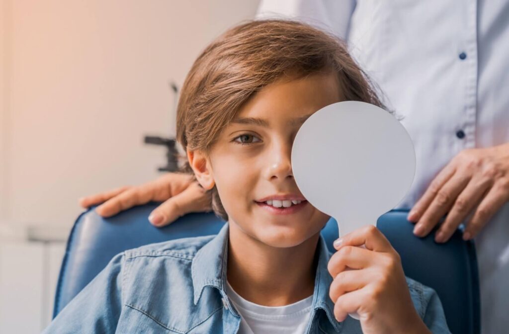 A young boy holding a white eye occluder over one eye during a vision test while an optometrist stands behind him.