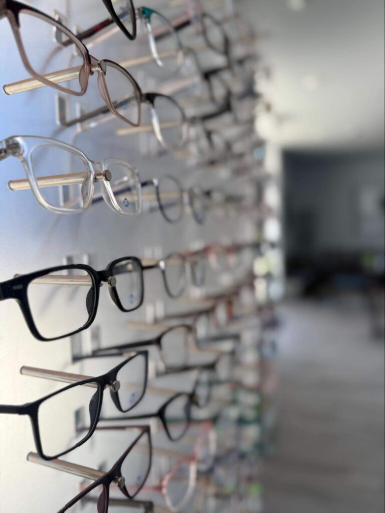 Eyeglasses displayed on a wall-mounted rack in an optical store, with various frames arranged in rows.